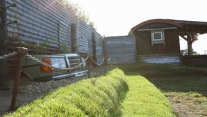 Une voiture ancienne est garée devant un bâtiment en métal ondulé auquel est adossée une petite structure en bois.