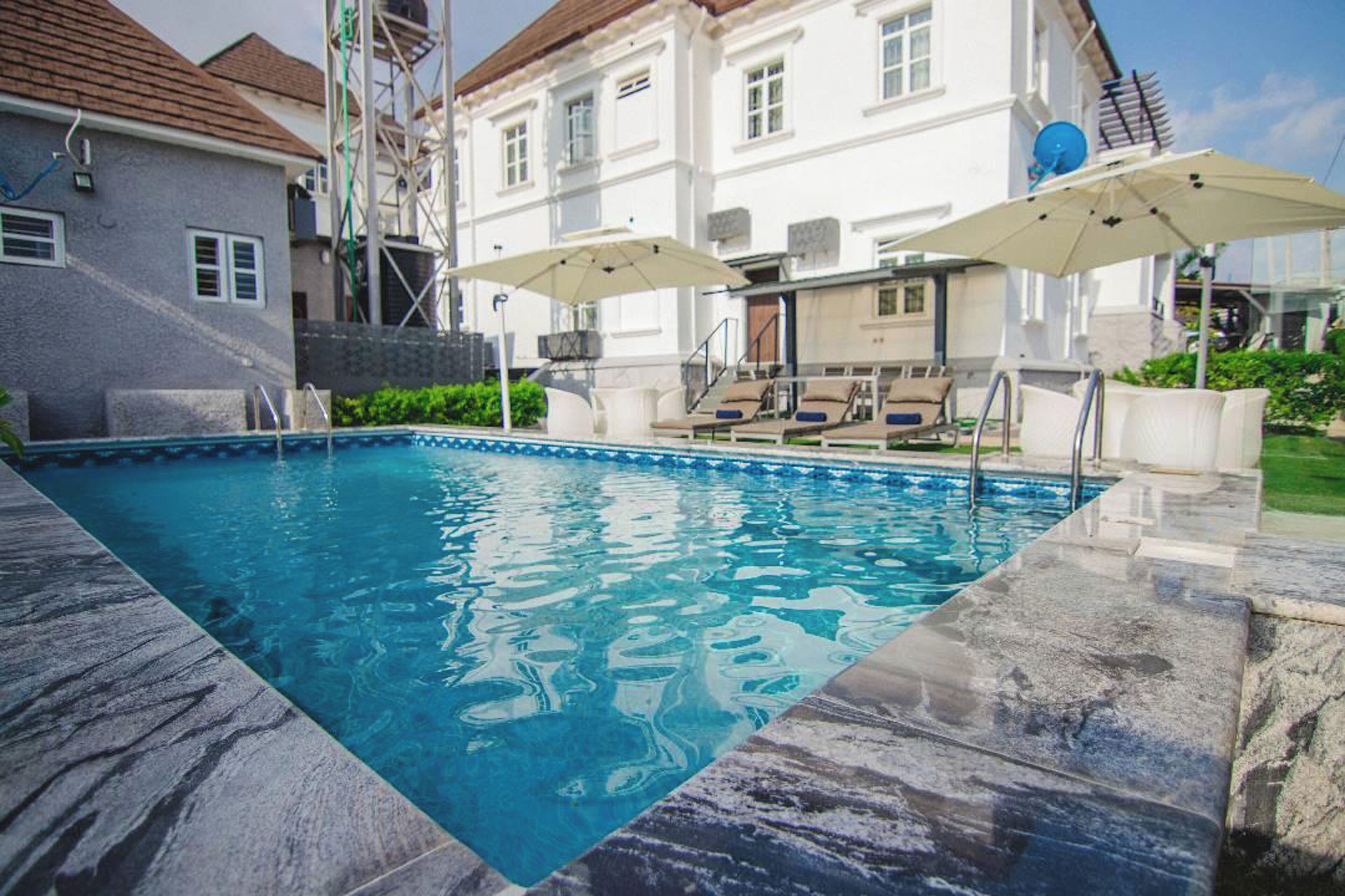 A swimming pool with a stone edge, two umbrellas, and a white building in the background.