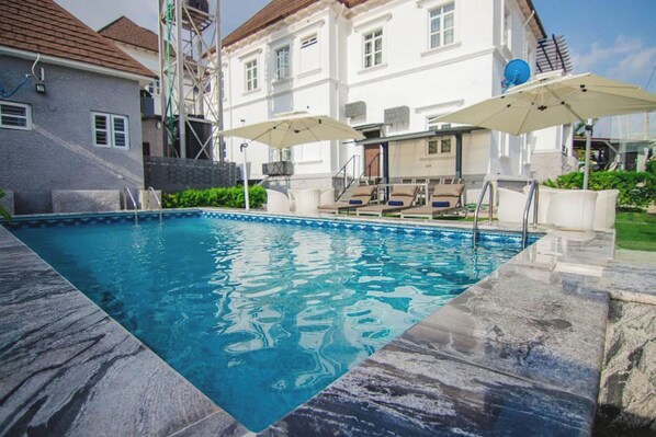 Ein Swimmingpool mit Steinkante, zwei Sonnenschirmen und ein weißes Gebäude im Hintergrund.