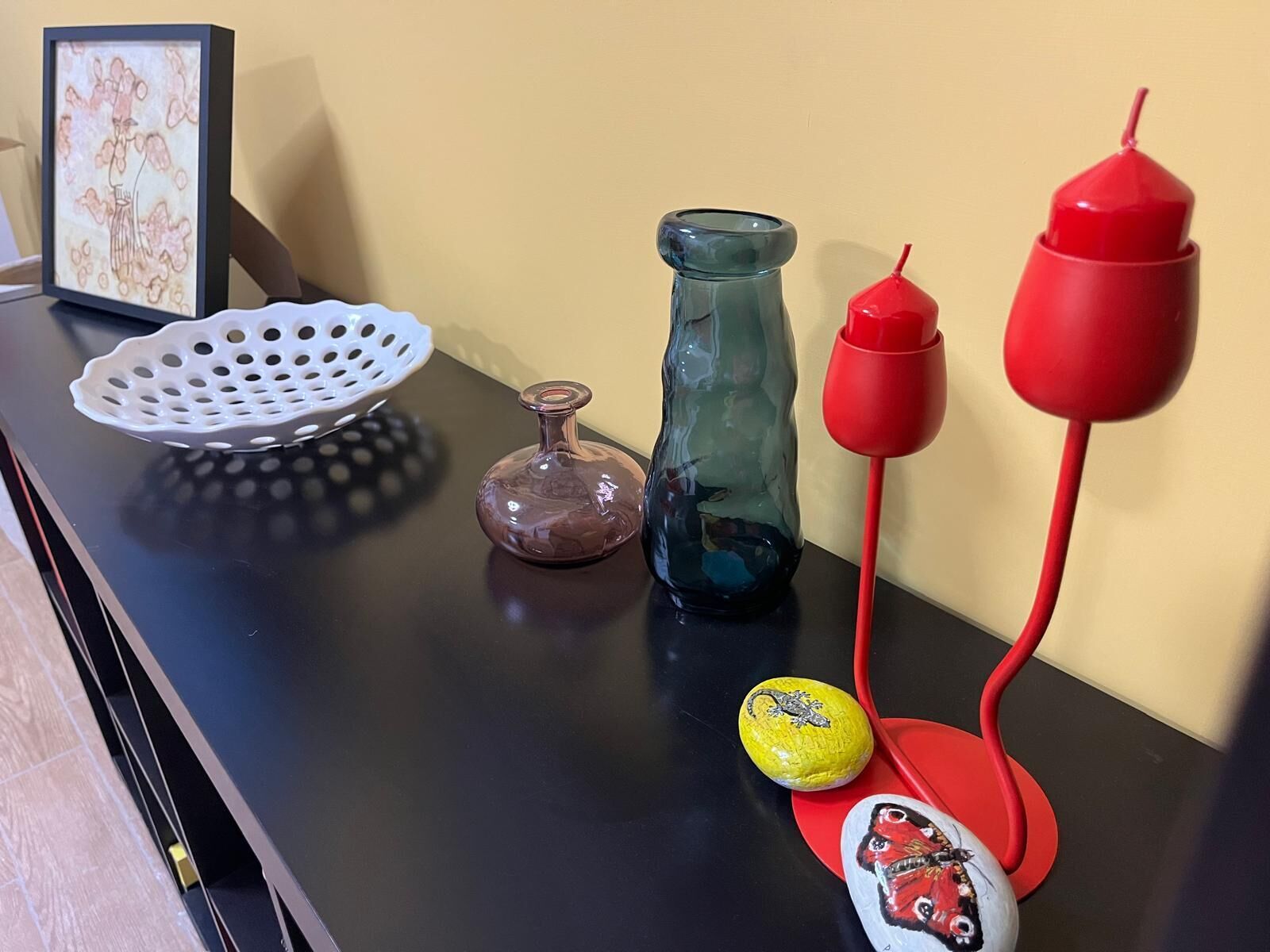 A table with a framed artwork, a glass vase, a ceramic pitcher, a ceramic bowl, and painted rocks.