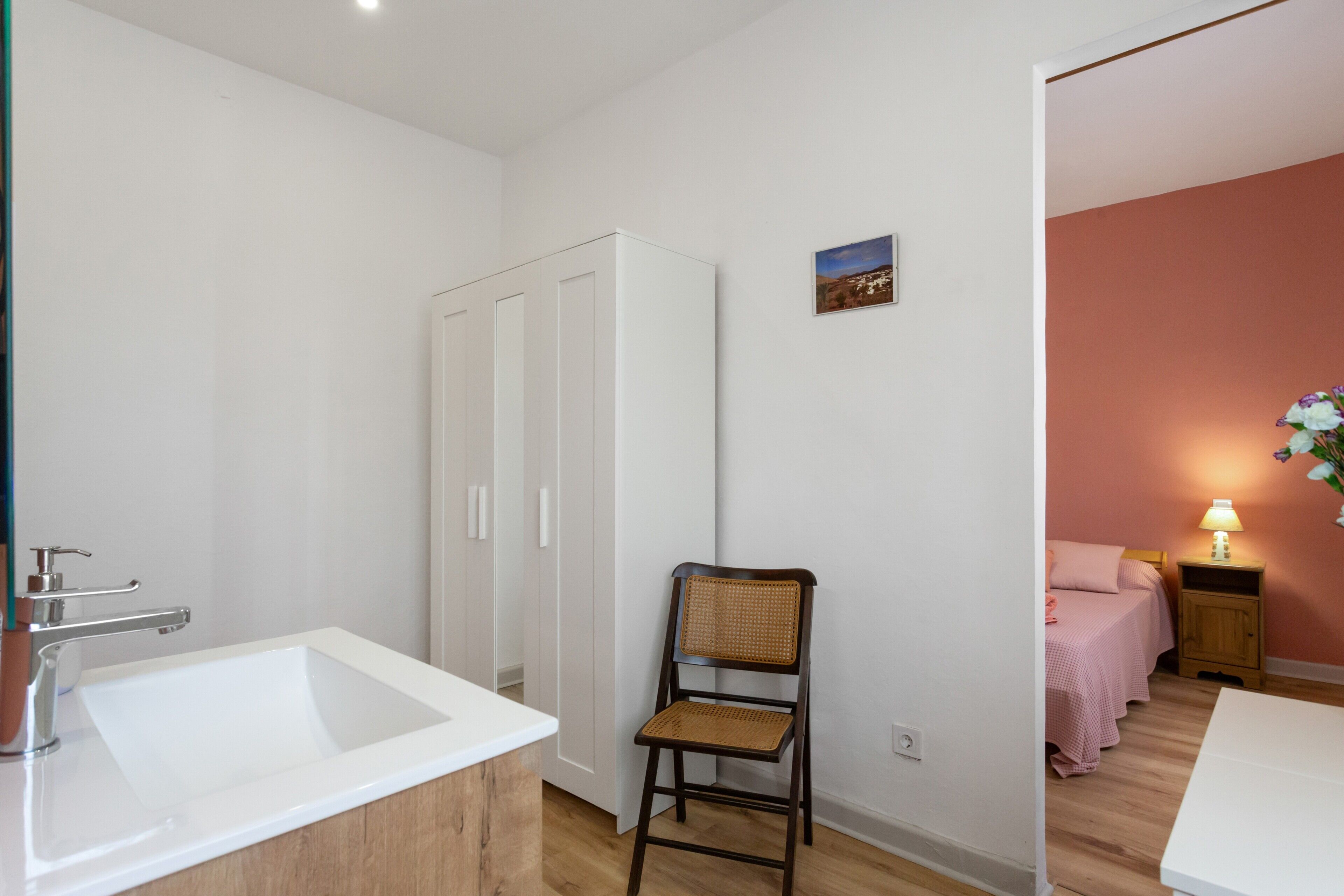 A bathroom with a white sink, a wooden chair, a wardrobe, and a bed with pink bedding.
