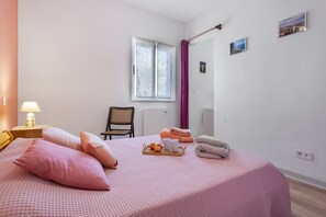 Een slaapkamer met een bed, kussens, een nachtkastje met een lamp, een stoel, een raam met gordijnen, en een muur met foto’s.