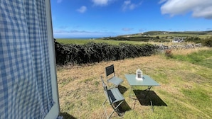 A view from a window overlooking a grassy field with a stone wall, a chair, and a table with a lantern. - Original Vintage Spirit Alpine Caravan Anglesey (Holyhead)