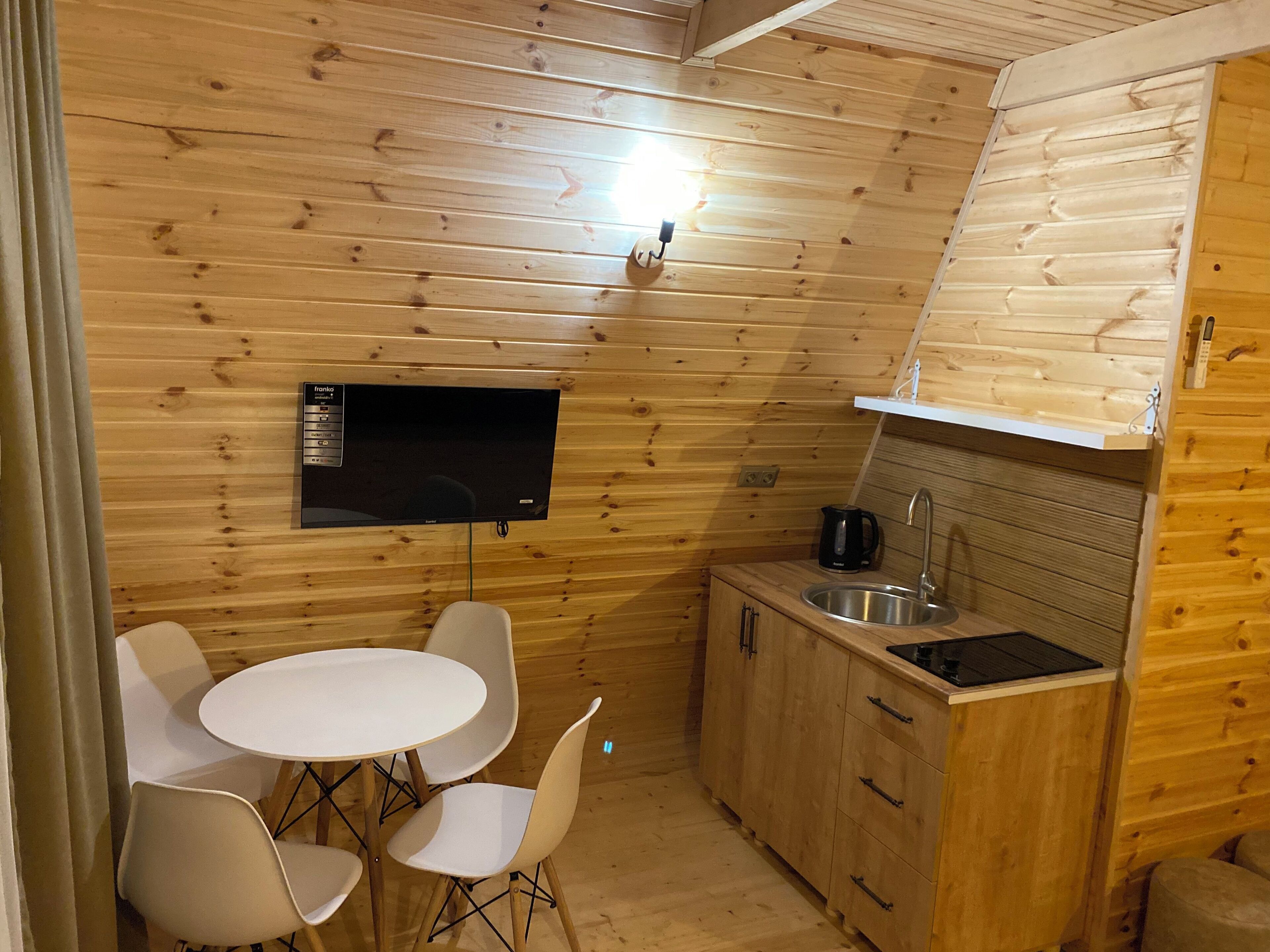Eine kompakte Küche mit Holzwänden, einem weißen Tisch mit Stühlen, einem schwarzen Wasserkocher und einem integrierten Fernseher.