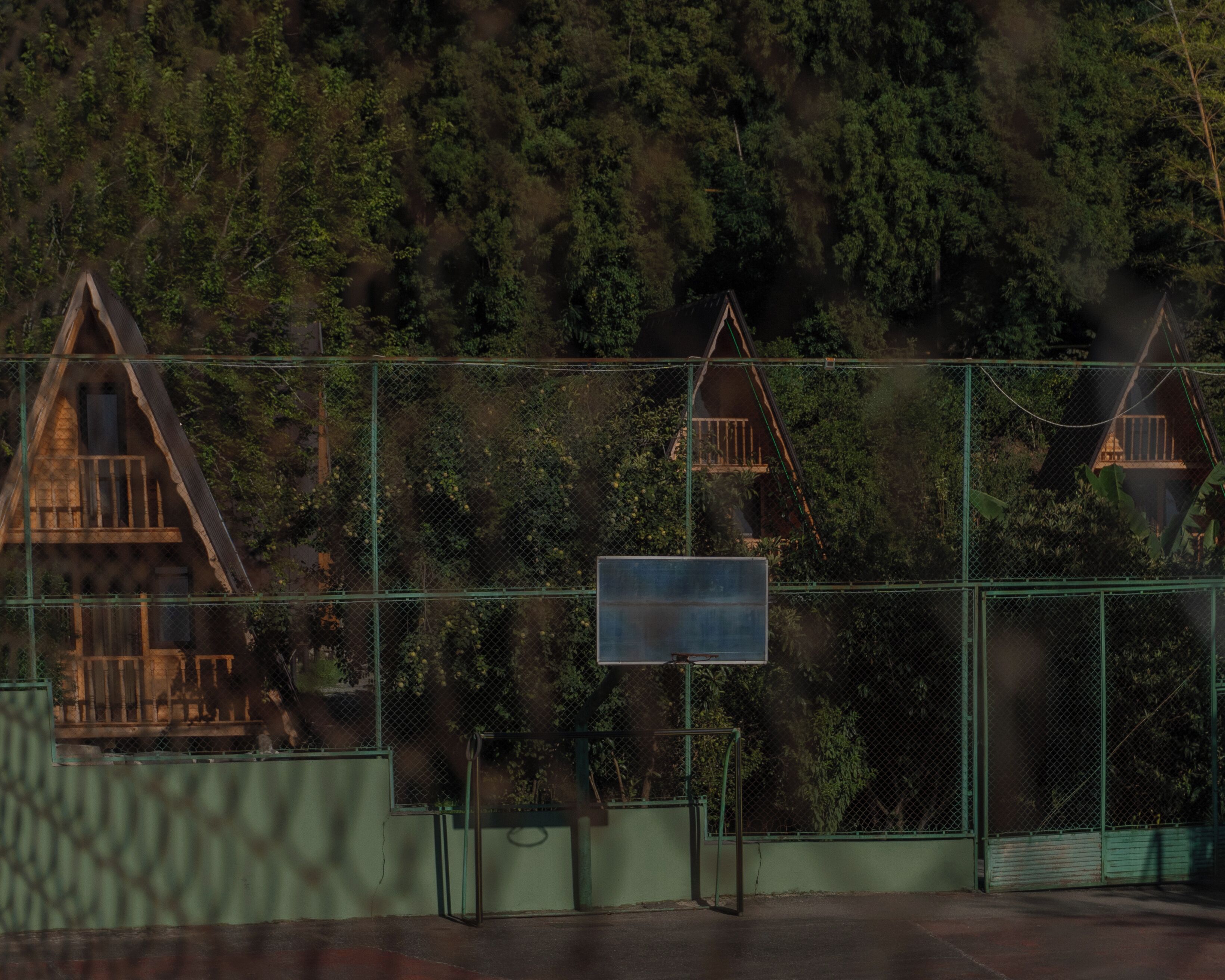 Un espacio cercado con una cancha de baloncesto, un cartel y un edificio con techo triangular al fondo.