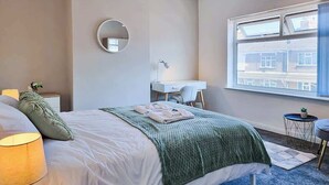 Ein Schlafzimmer mit Bett, Nachttischlampe, rundem Spiegel, Schreibtisch, Stuhl und einem Fenster mit Blick auf Gebäude.