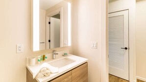 Une salle de bain avec un lavabo blanc, un miroir et un meuble en bois. Il y a une serviette sur le comptoir et une porte avec une fenêtre.