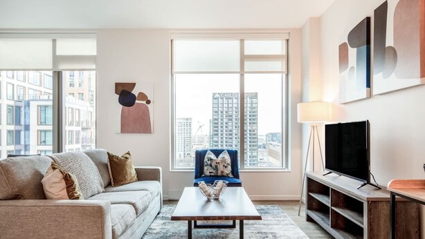 A modern living room with a sofa, a coffee table, a TV, and a large window with a city view. - Luxury Apt in Seaport with Gym and Pool (Boston)