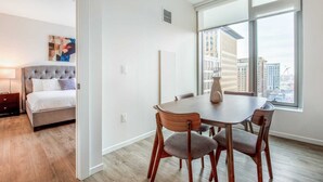 A modern hotel room with a large window, a dining area with a table and chairs, a bed with a tufted headboard, and a painting on the wall. - Luxury Apt in Seaport with Gym and Pool (Boston)