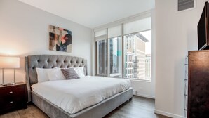 A modern bedroom with a large bed, a nightstand, a window with a city view, and a piece of abstract art on the wall. - Luxury Apt in Seaport with Gym and Pool (Boston)
