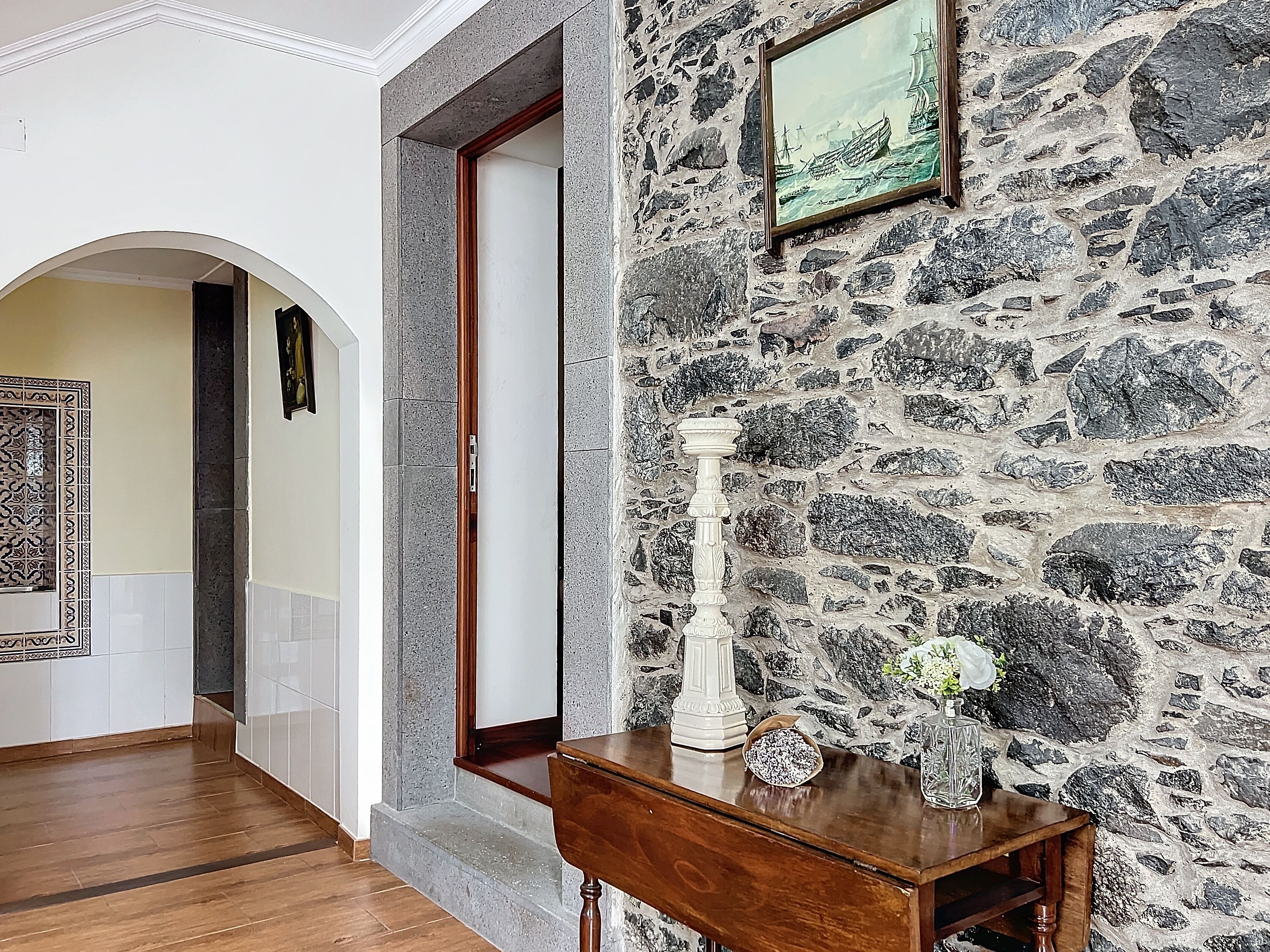 A hallway with a stone wall, a wooden table with a vase and flowers, and a framed picture on the wall.