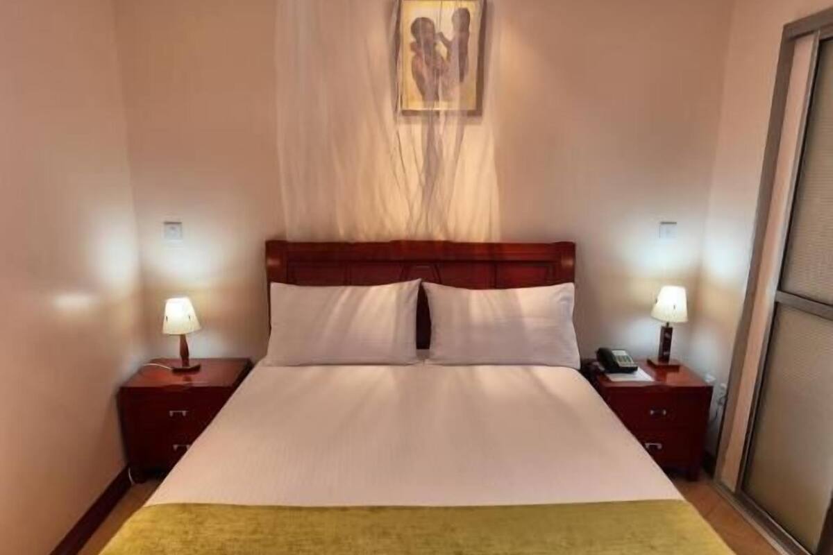 A neatly made bed with a green throw blanket, two bedside tables with lamps, a large painting on the wall, and a sliding door on the right.