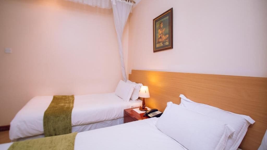 A hotel room with two beds, a bedside table with a lamp, a phone, and a picture on the wall.