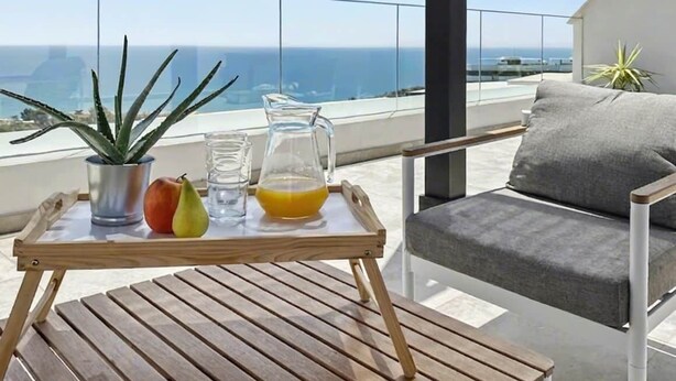 A wooden table with a tray holding a pitcher, glasses, and fruits, set on a balcony with a view of the ocean.