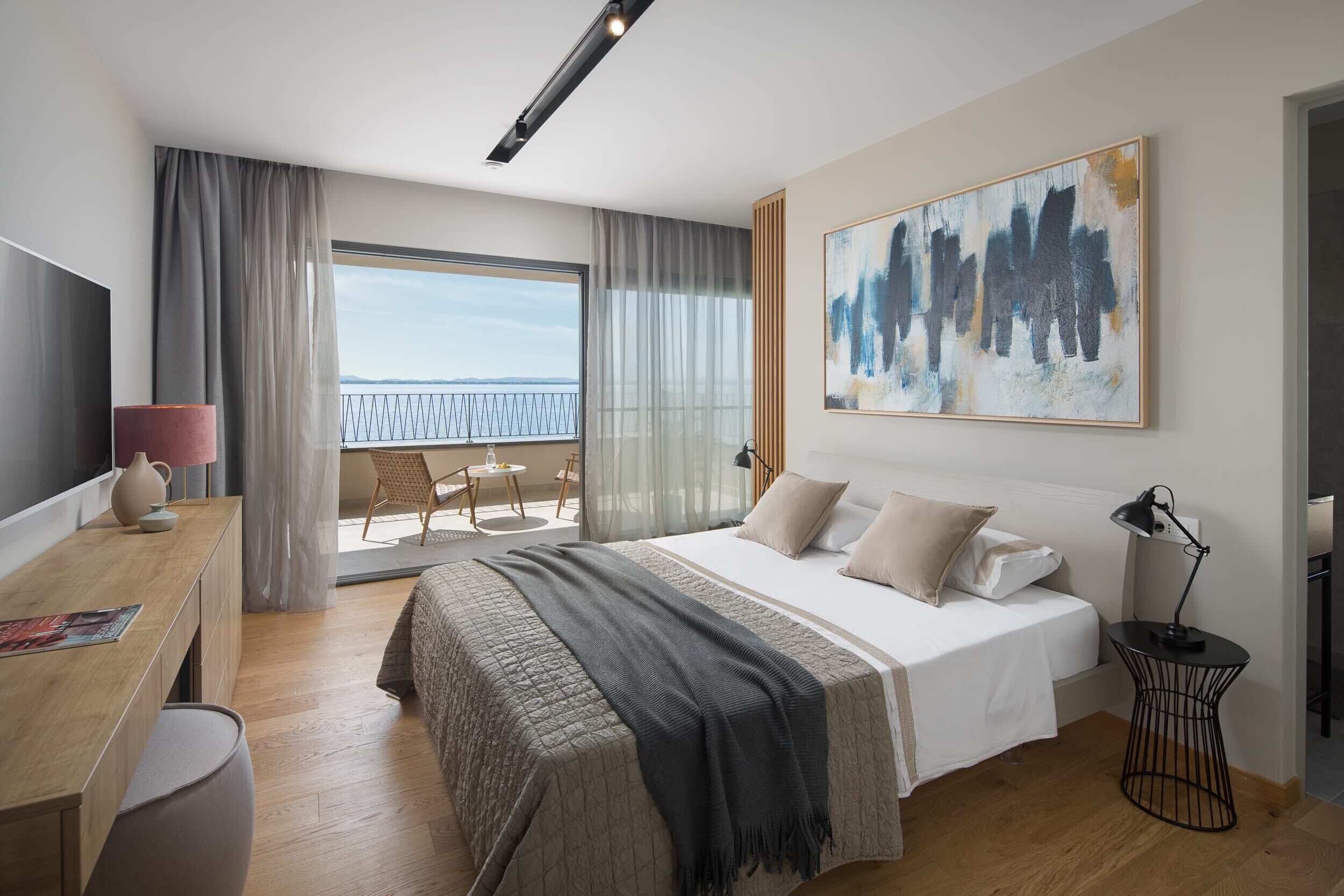 Ett modernt hotellrum med en stor säng, ett skrivbord, en stol, en TV och en balkong med utsikt över havet.