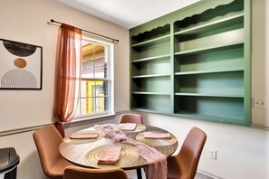 A dining area with a round table, chairs, a window with curtains, and a green shelf unit.
