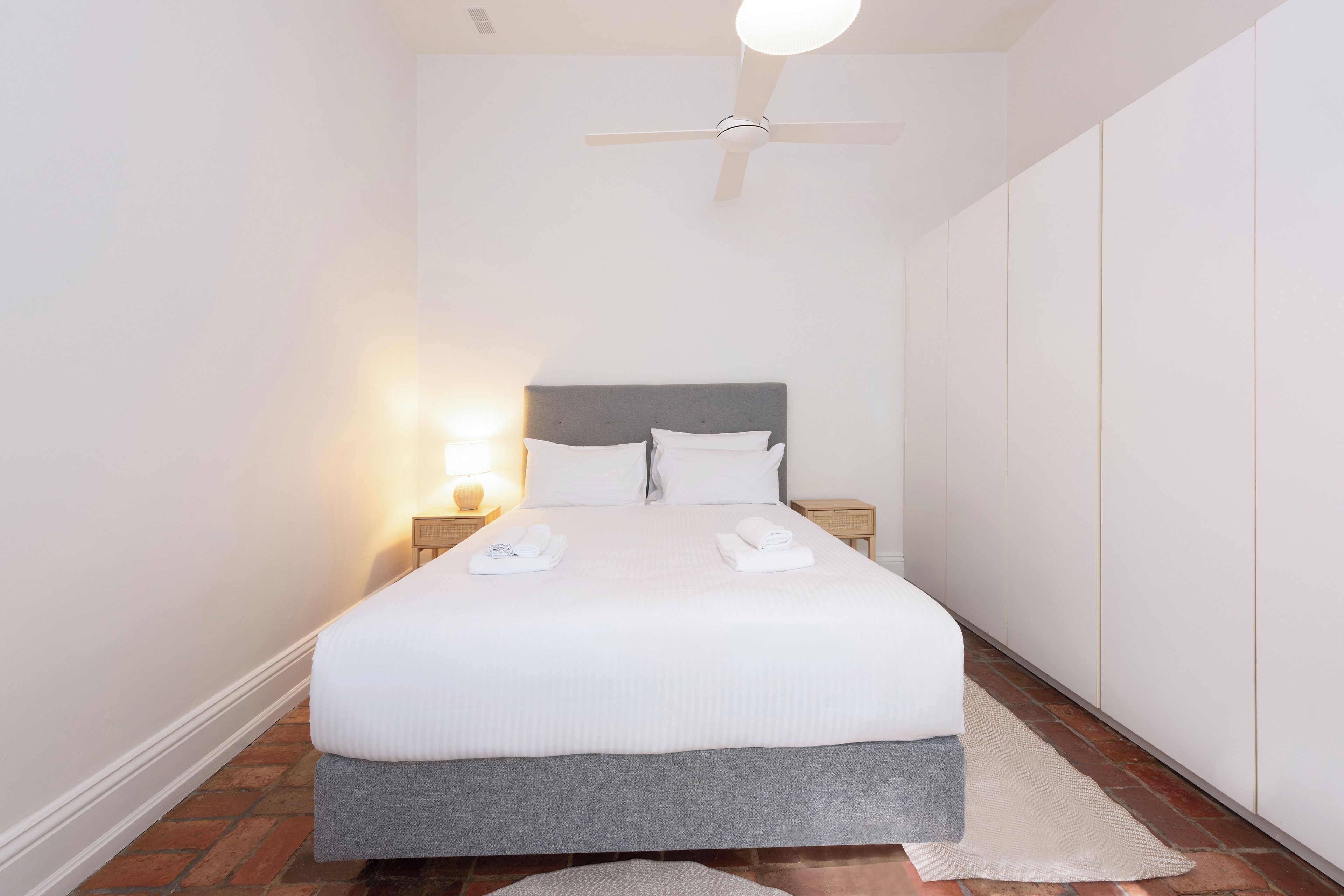 A neatly made bed with white linens and a grey headboard in a room with white walls and a ceiling fan.