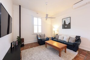 Ein Wohnzimmer mit einem schwarzen Ledersofa, einem Holztisch, einem gemusterten Teppich, einem Deckenventilator und einem gerahmten Bild an der Wand.