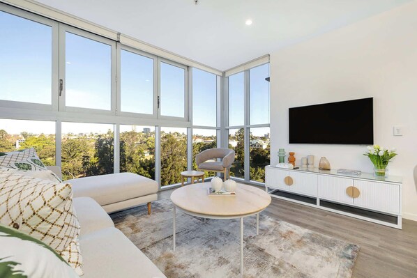 A modern living room with a large window, a sofa, a round table, and a TV mounted on the wall.