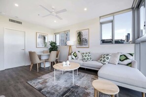 A modern living room with a sofa, a round coffee table, a dining table with chairs, a ceiling fan, and large windows with a city view. - 2BR South Brisbane | Free Parking + Walk to QPAC (South Brisbane)