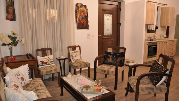 A living room with a sofa, chairs, a coffee table, and a kitchen area with wooden cabinets and a window. - Holiday Home 'House For Rent In Apostolous' with Mountain View, Private Garden and Wi-Fi (Iraklio)