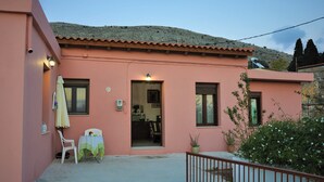 A pink building with a door, windows, a small table with a chair, and a potted plant. - Holiday Home 'House For Rent In Apostolous' with Mountain View, Private Garden and Wi-Fi (Iraklio)