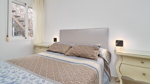 A bedroom with a bed, two bedside tables, a window with curtains, a wall-mounted light, and a staircase visible through the window.