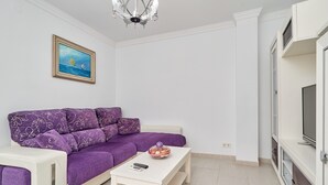 A modern living room with a purple sofa, a white coffee table, a painting on the wall, and a television on a cabinet.