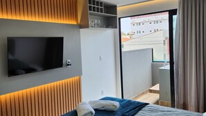A modern hotel room with a bed, a flat-screen TV mounted on the wall, a wardrobe, and a balcony with a view of the city. - Flat Ofurô 15 VP by La Playa (Ipojuca)