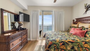 A bedroom with a bed, a dresser, a television, a balcony with curtains, and a view of the ocean. - Emerald Isle 1709 - Beachfront condo! Front row beach chairs included! (Panama City Beach)