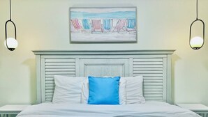 Un lit bien fait, avec du linge de lit blanc et un seul oreiller bleu, une tête de lit de couleur claire et, au-dessus, un tableau représentant des chaises de plage.