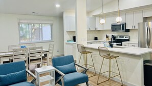 Une cuisine moderne avec des meubles blancs, un îlot central doté de tabourets de bar et un coin repas avec une table en bois et des chaises bleues.