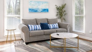 A living room with a grey sofa, a round coffee table, and a painting on the wall. - Spacious Raleigh Retreat | Near Downtown + I-440 (Raleigh)