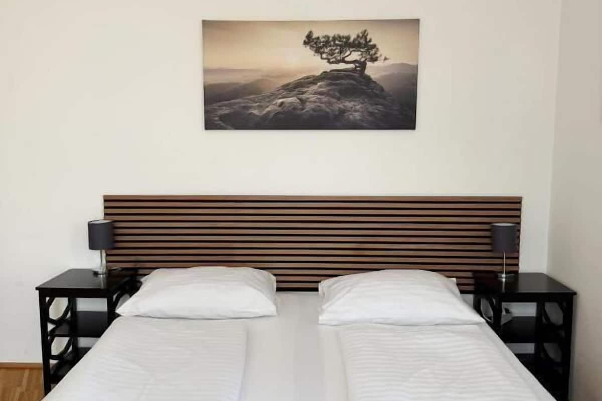 A double bed with white linens, a wooden headboard, two bedside tables with lamps, and a framed picture of a tree on a mountain.