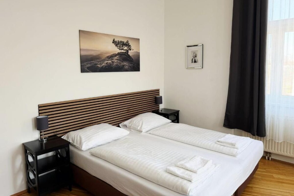 A double bed with white linens, a wooden headboard, two bedside tables with lamps, a framed picture on the wall, and a window with curtains.