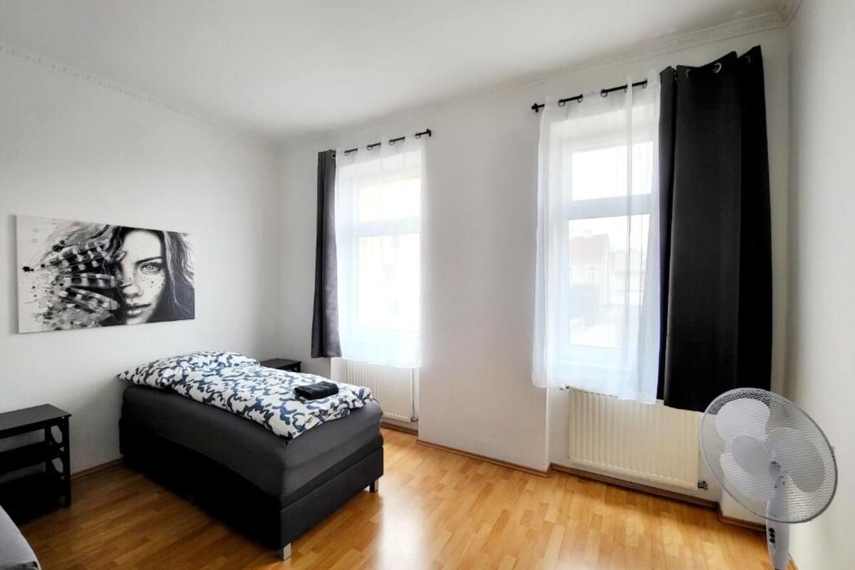 Une chambre à coucher moderne avec un lit, une table de chevet, un ventilateur sur pied et une fenêtre avec des rideaux.