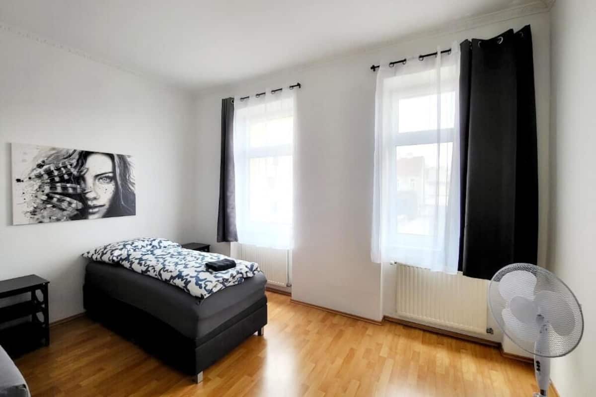 A modern bedroom with a bed, a bedside table, a standing fan, and a window with curtains.