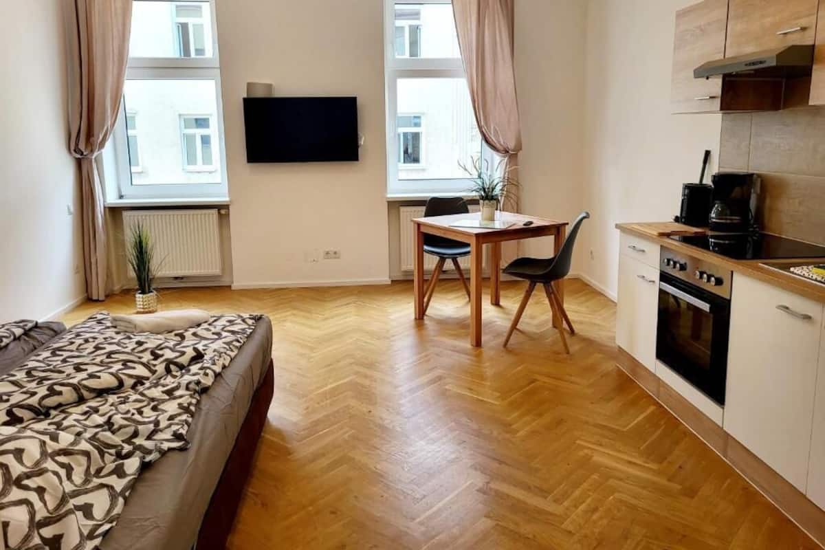 A compact living space with a bed, a dining area with a table and chairs, a kitchenette, and a TV mounted on the wall.