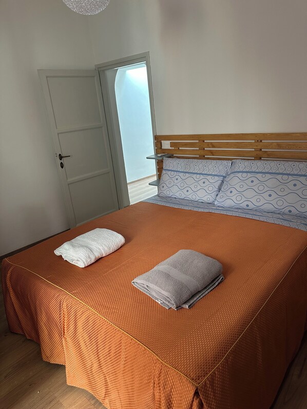 A neatly made bed with an orange bedspread, two folded towels, and a wooden headboard with patterned pillows.