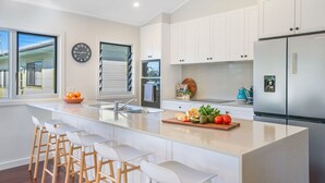 A modern kitchen with a large island, white cabinetry, stainless steel appliances, and a view of the outdoors. - Luxury Waterfront Home (Evans Head)