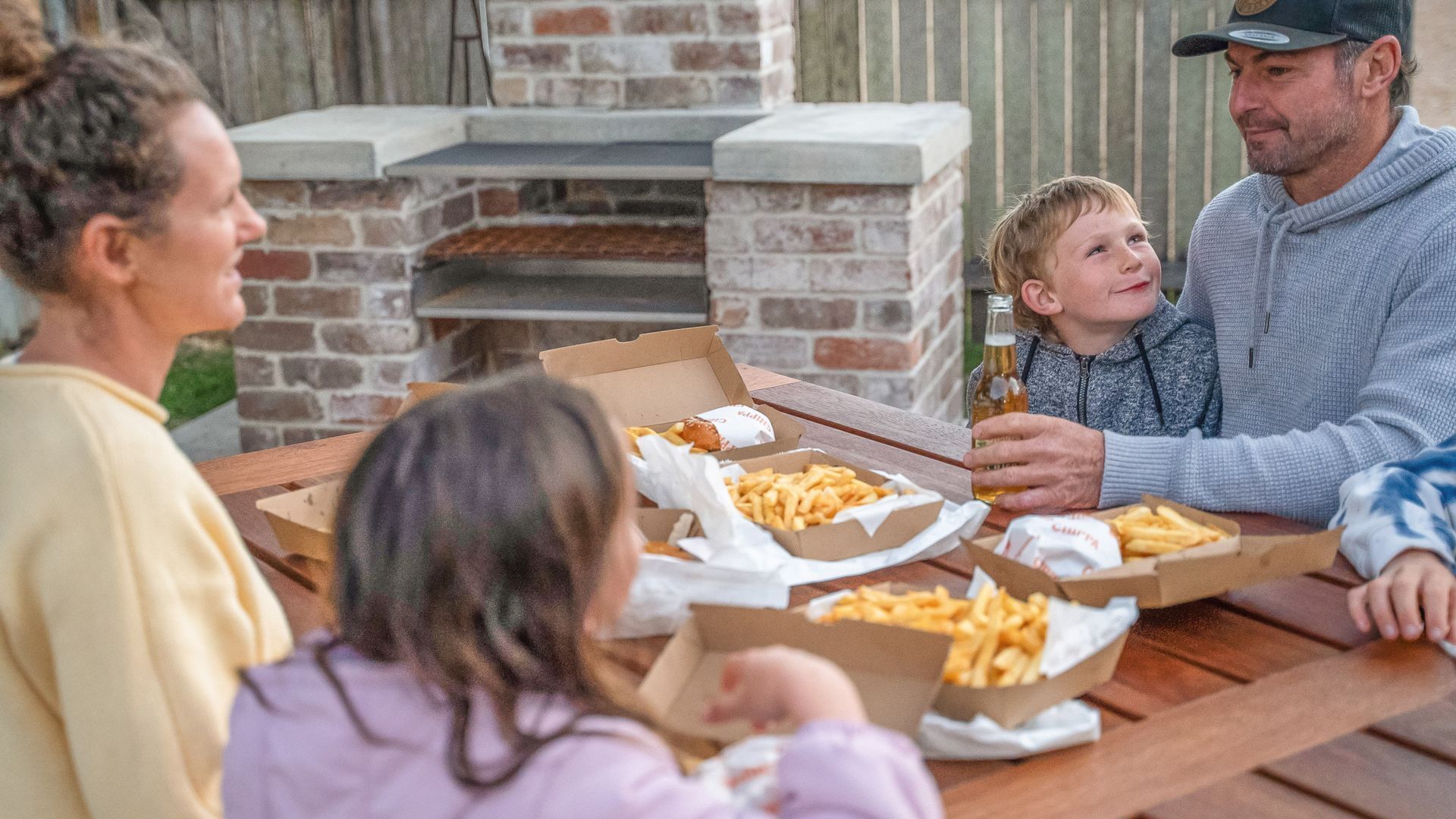 Eine Familie isst an einem Tisch im Freien mit Pizzakartons und Pommes, vor einem Kamin aus Ziegelsteinen.