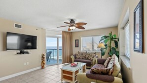 A living room with a flat-screen TV, a ceiling fan, a sofa, a coffee table, and a large window with blinds. - Spacious 2 bedroom unit overlooking the ocean! | Blue Water Keyes -  1108 (North Myrtle Beach)