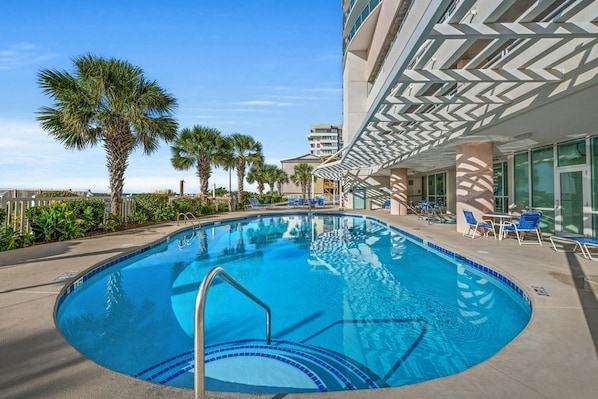 A swimming pool with a poolside area, palm trees, and a modern building with a geometric facade.