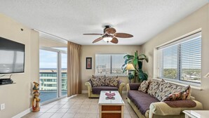 A living room with a ceiling fan, a flat-screen TV, a sofa, a coffee table, and a large window with blinds. - Spacious 2 bedroom unit overlooking the ocean! | Blue Water Keyes -  1108 (North Myrtle Beach)