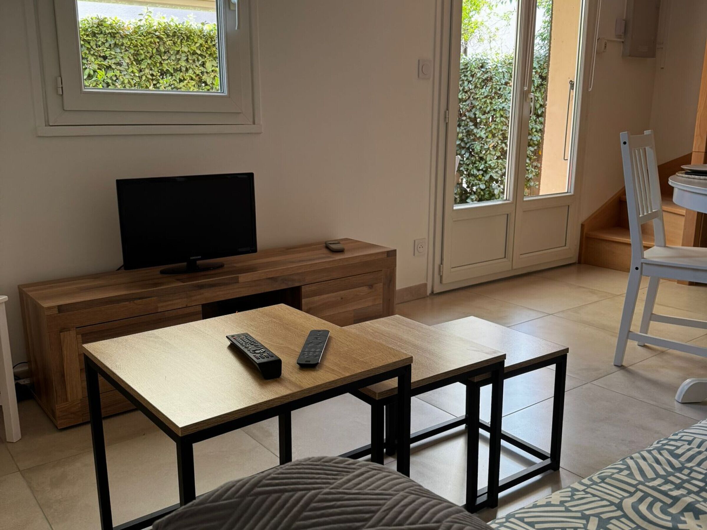 Habitación con una televisión sobre un mueble de madera, una mesa con dos mandos a distancia, una silla blanca y vistas a través de un ventanal.