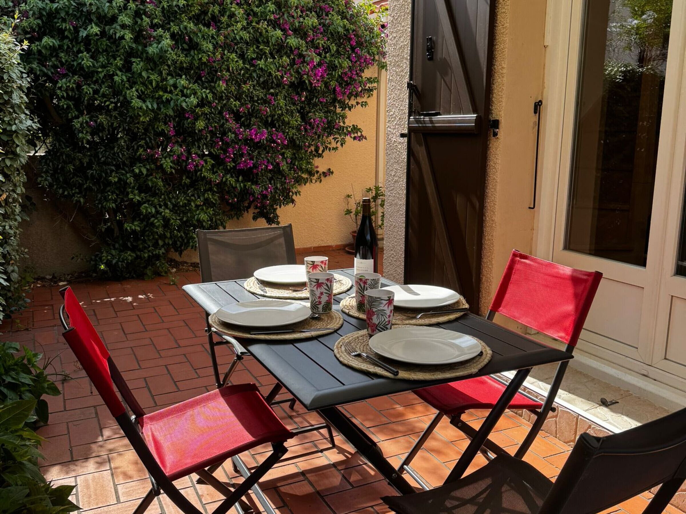 Un patio esterno con un tavolo apparecchiato per due, circondato da sedie rosse, una pianta in vaso e una porta.