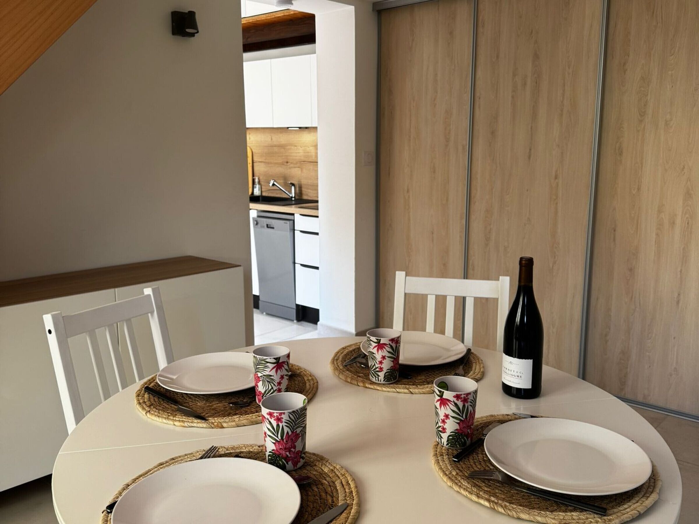 A dining table set with plates, cutlery, and floral-patterned cups, with a bottle of wine on the table.