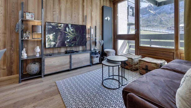 Ein modernes Wohnzimmer mit großem Fernseher, Holzwandverkleidung, einer gemütlichen Couch und Bergblick durch große Fenster.