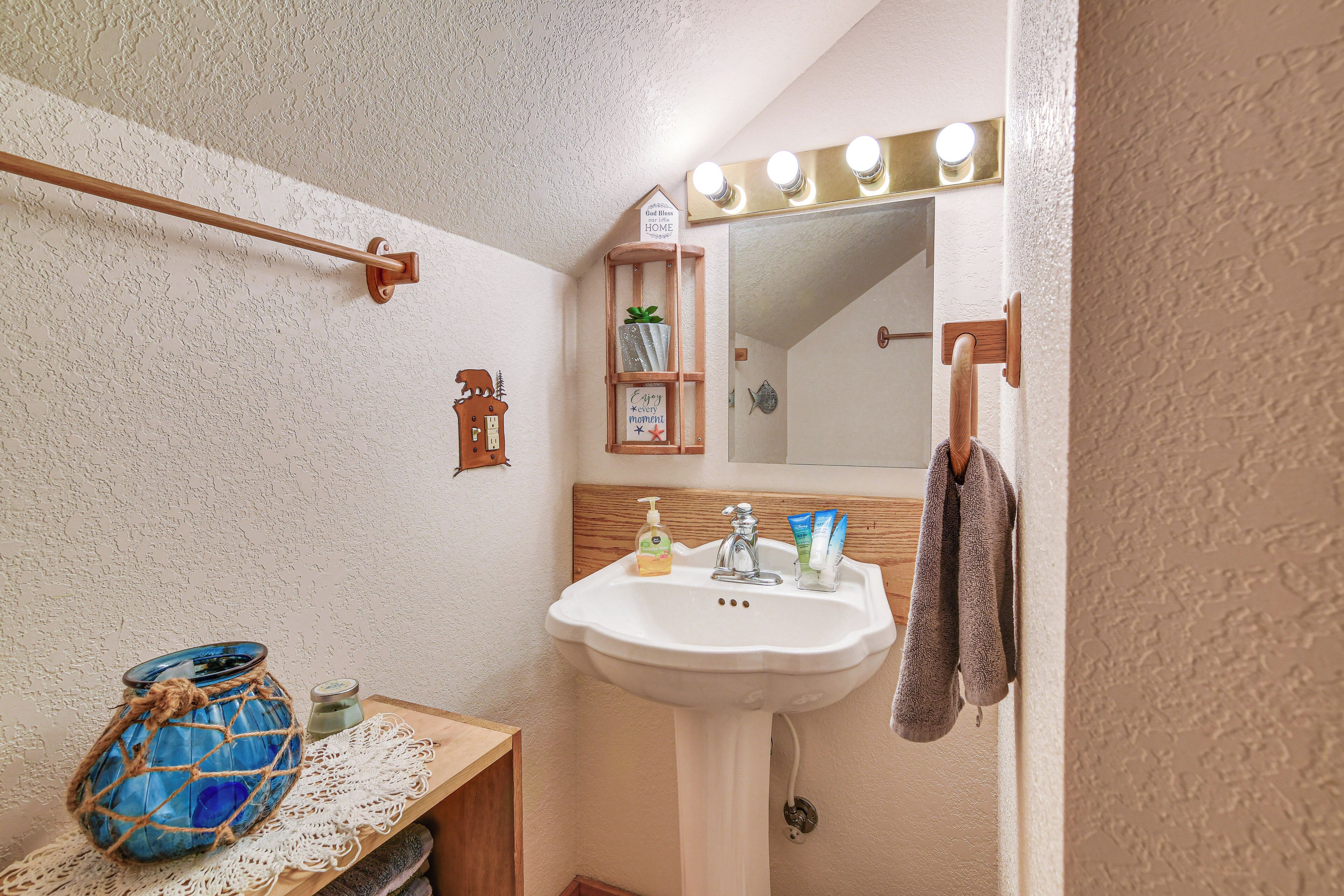 Un baño con un lavamanos blanco, un estante de madera con artículos de tocador, un florero azul y un espejo con luces arriba.