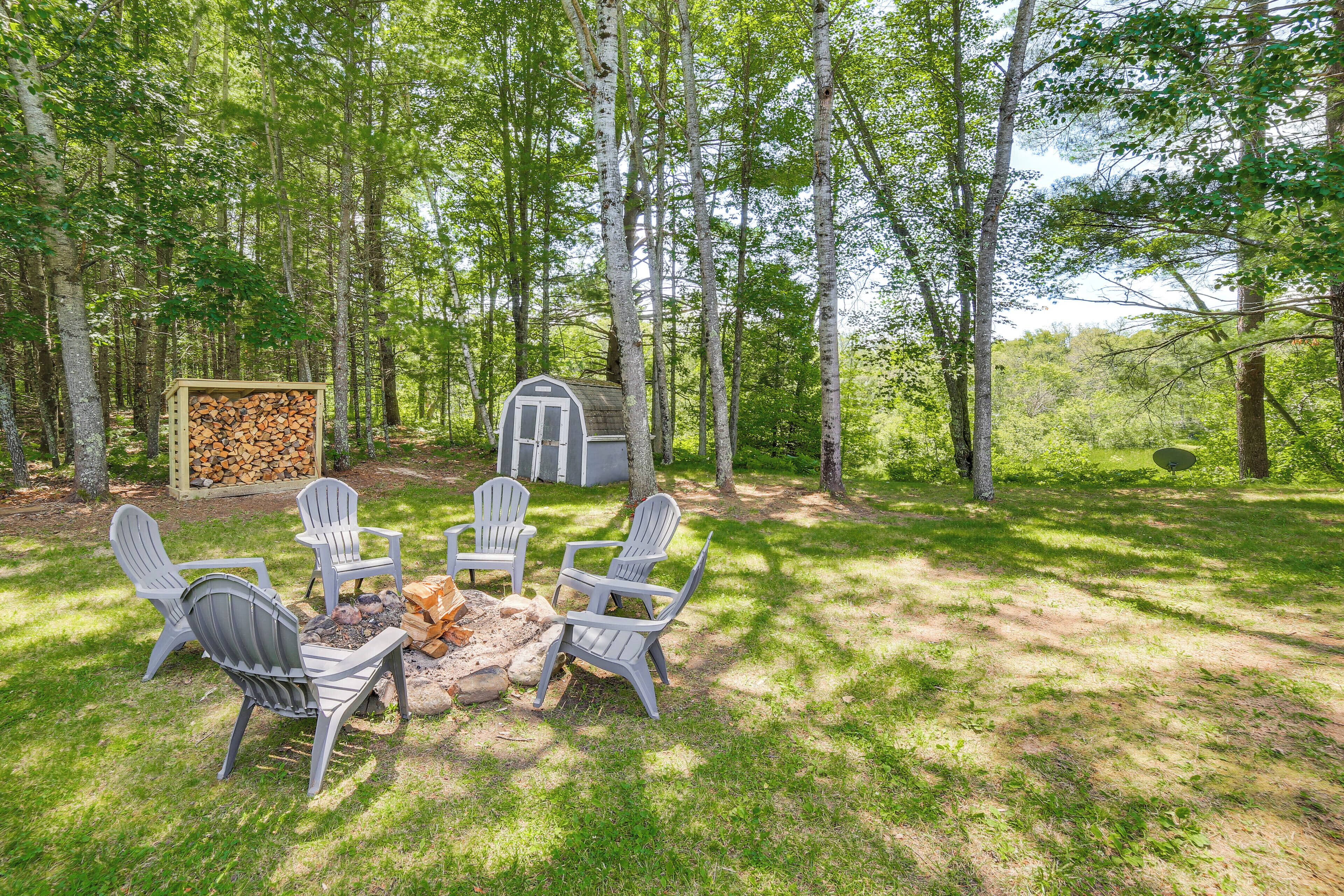 A forested area with a fire pit and Adirondack chairs.
